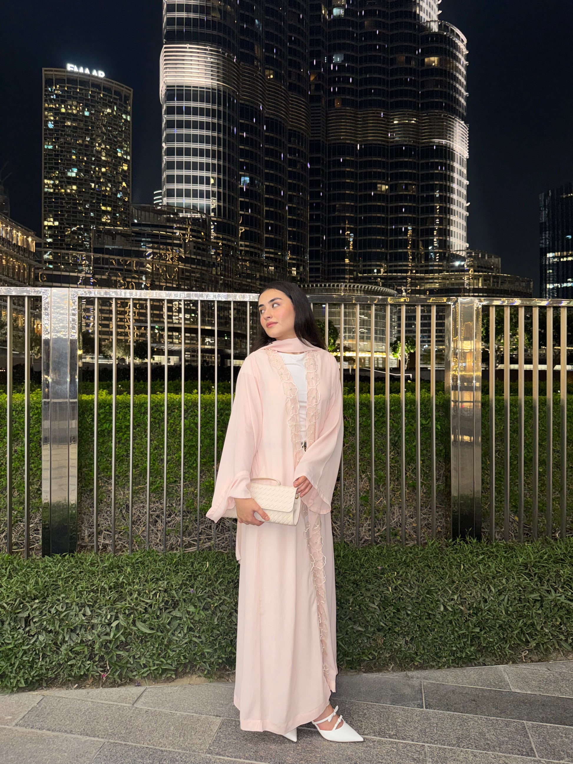 A woman wearing a light pink traditional long robe and a matching headscarf stands outdoors at night in front of a metal fence and illuminated skyscrapers. She is holding a small cream-colored clutch and wearing white pointed flats.