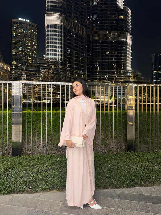 A woman wearing a light pink traditional long robe and a matching headscarf stands outdoors at night in front of a metal fence and illuminated skyscrapers. She is holding a small cream-colored clutch and wearing white pointed flats.