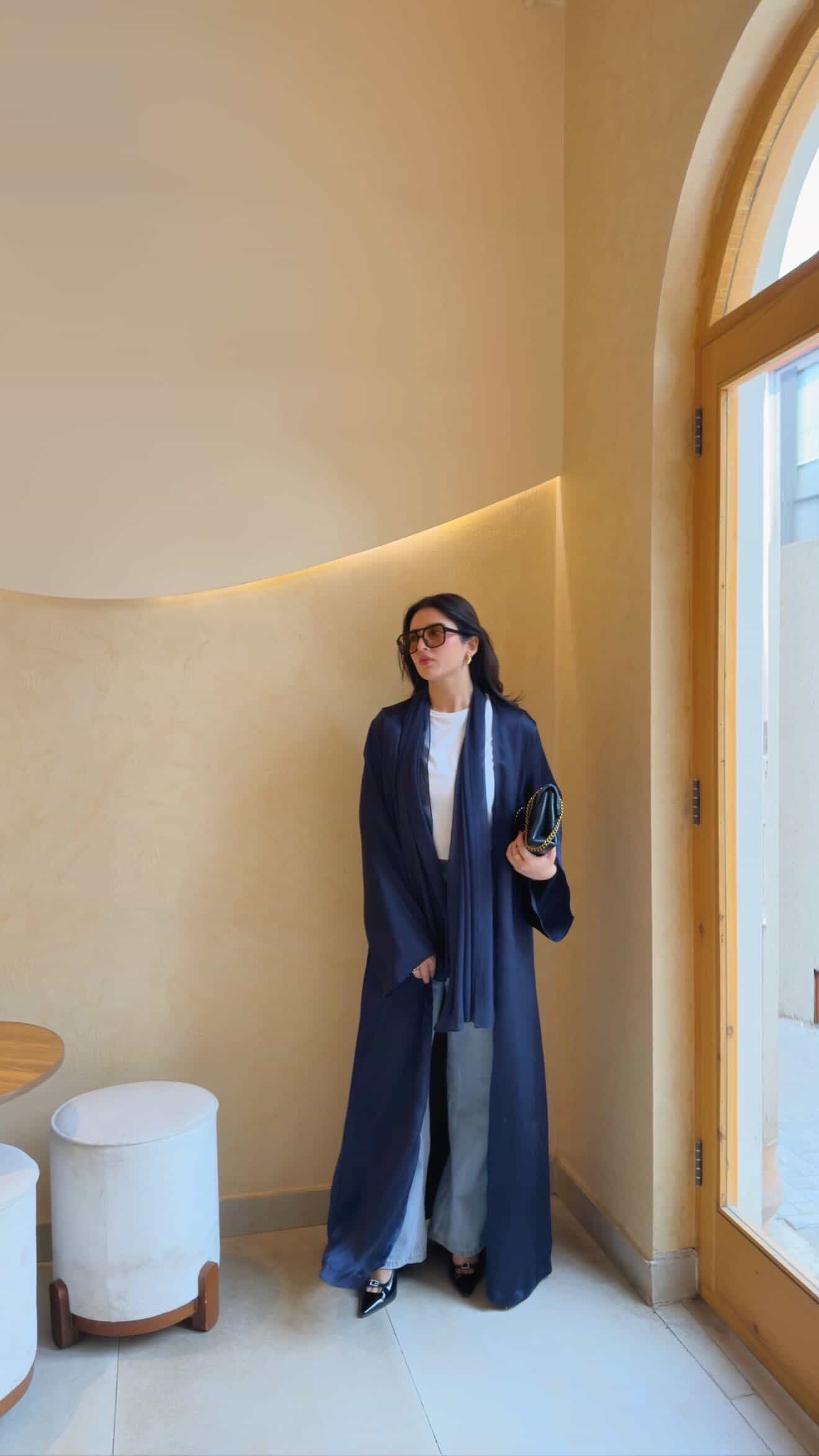 A woman wearing a long navy blue open-front abaya with a matching scarf, layered over a white top and gray pants, standing in a warm beige interior hallway.