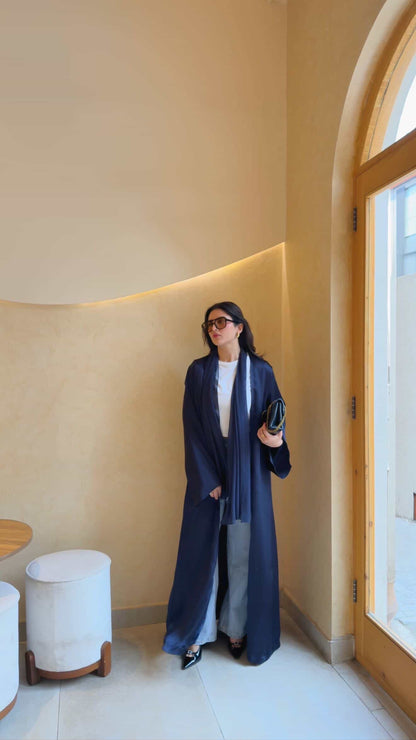 A woman wearing a long navy blue open-front abaya with a matching scarf, layered over a white top and gray pants, standing in a warm beige interior hallway.